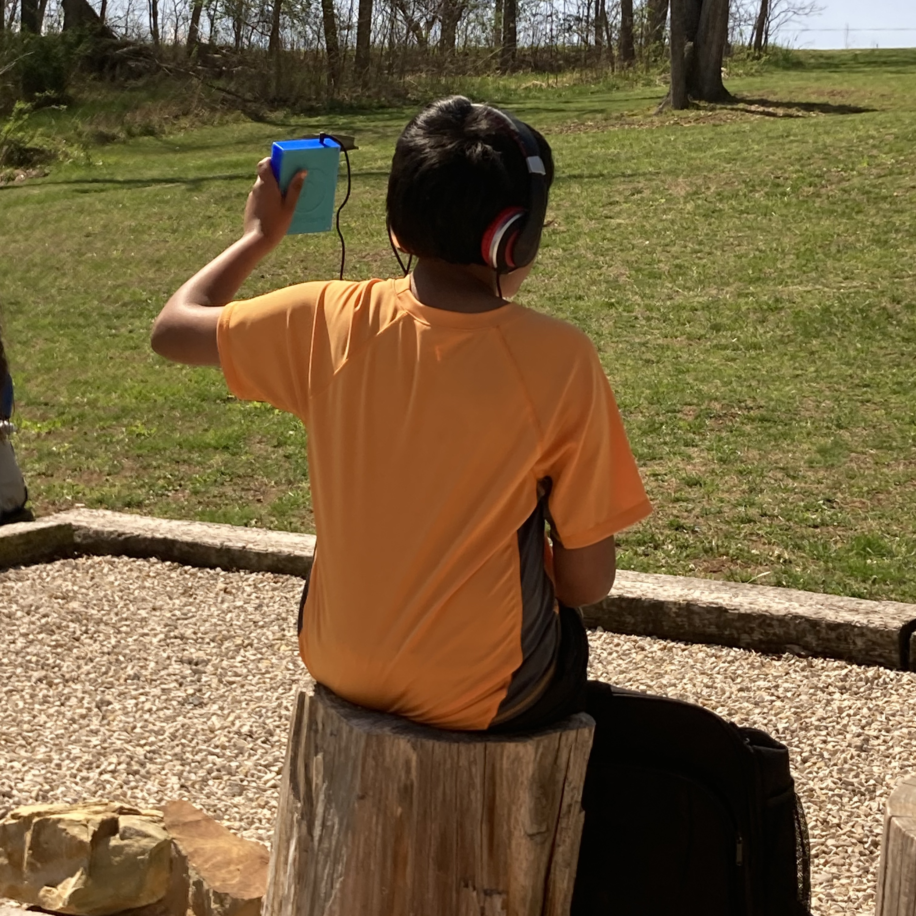 A young boy sitting on a tree stump wearing headphones and holding a LightSound device pointed at the sun.