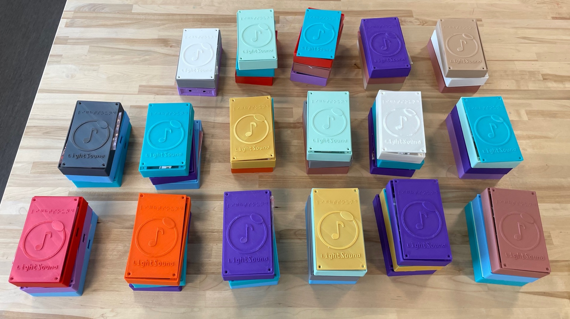 A table with three rows of 3D printed LightSound device cases. The cases are all different colors and stacked about three devices high. The case is about the size of a smart phone and the lid has the LightSound logo which is a musical note in a circle with the word LightSound underneath and LightSound written in Braille above.