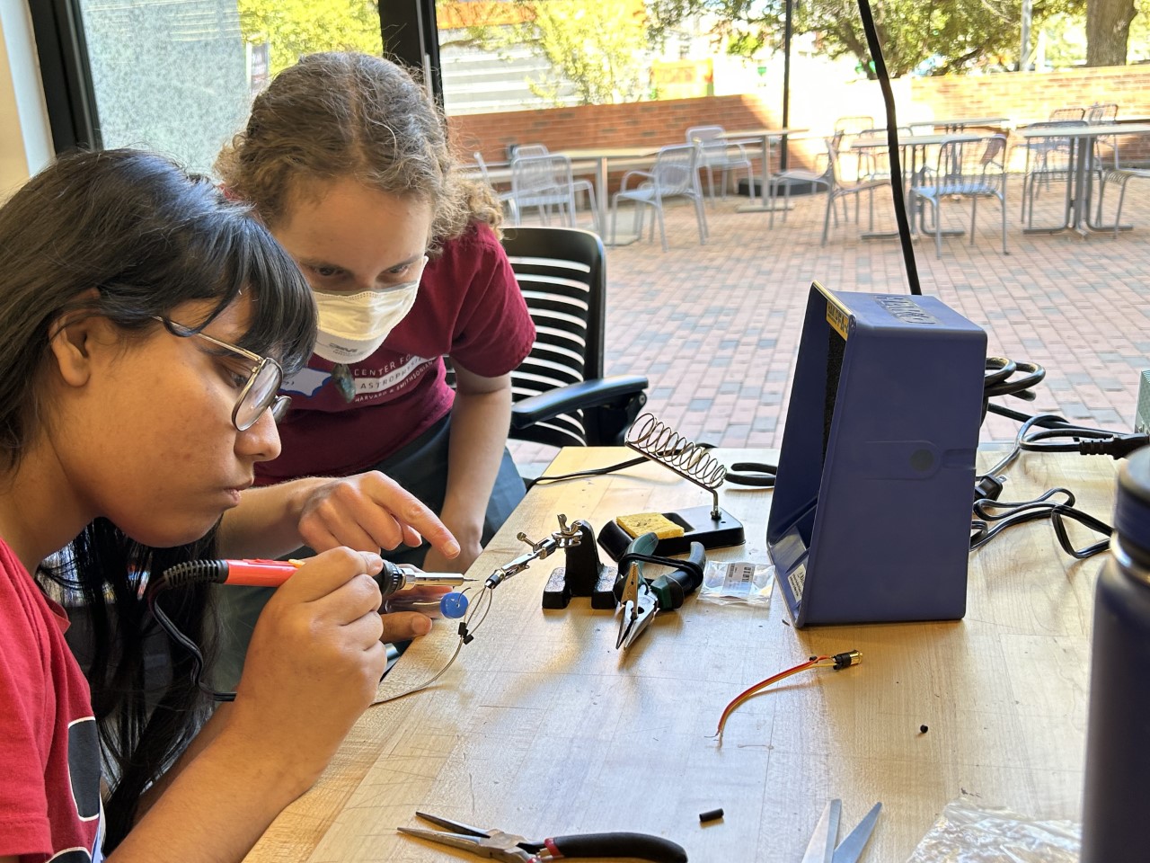 A woman with dirty blond, curly hair in a ponytail pointing to a LightSound component while teaching a female student how to solder at a University of Arizona LightSound workshop.